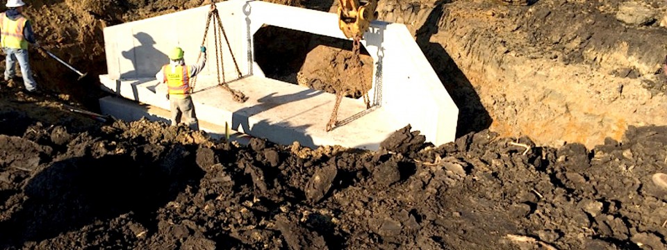 Construction workers installing a large precast concrete drainage structure in an excavated trench using a crane